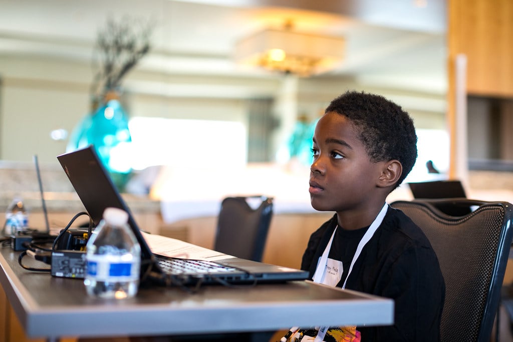 A black child sits at a desk with an open laptop and a bottle of water.