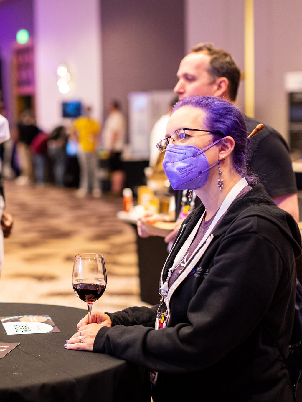 Woman with purple hair, glasses, and a purple mask leans agains a table with a glass of red wine.