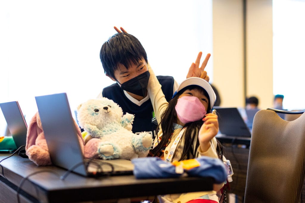Two Asian kids wearing masks sit in front of computers while making bunny ears with their fingers.