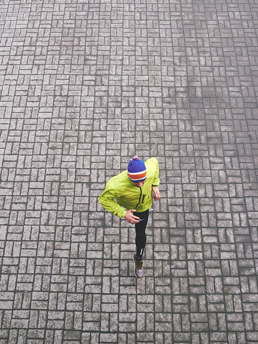 A person wearing a bright yellow jacket and blue hat runs across the brick pavement.
