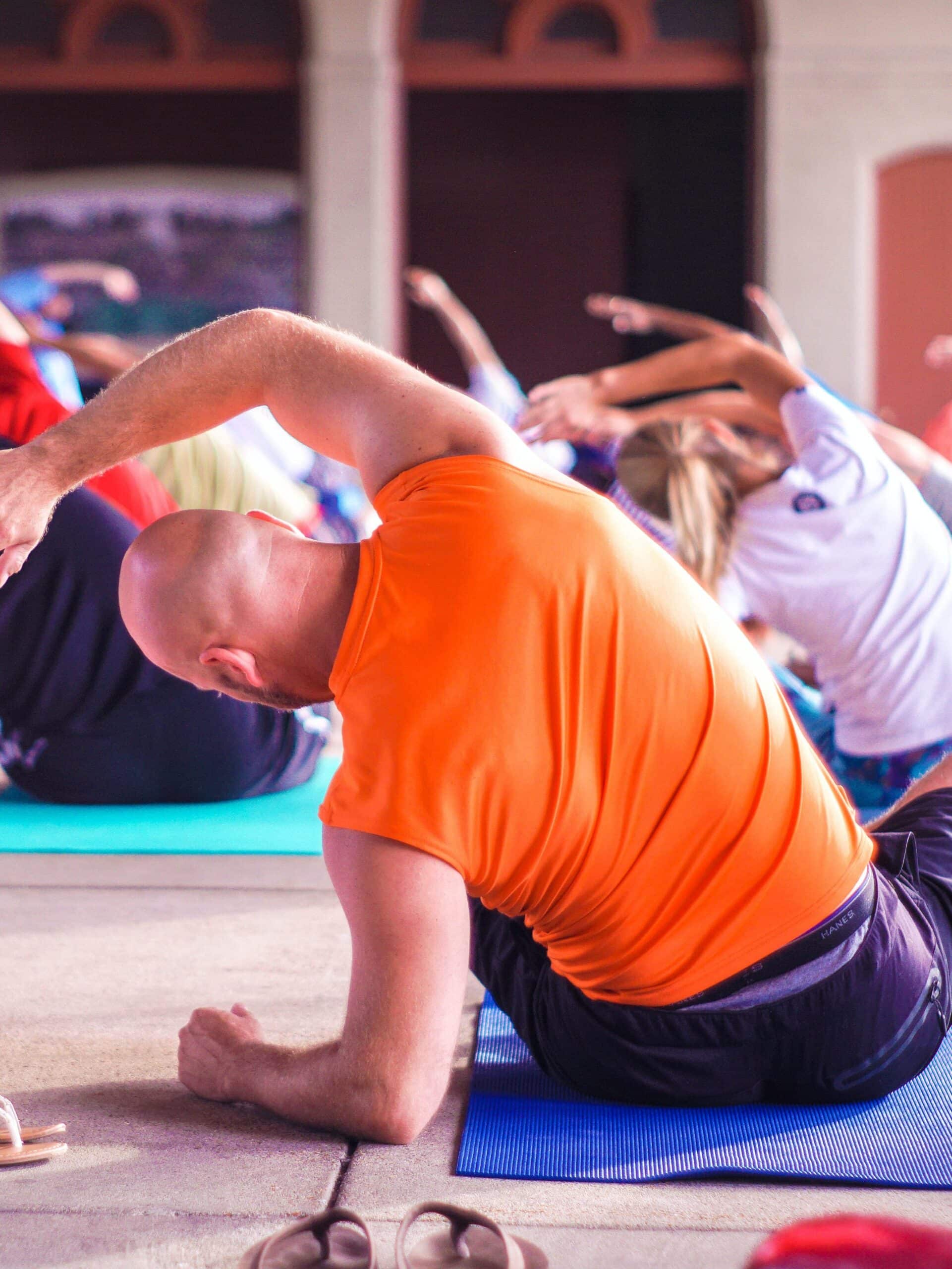 Bald man in orange tank top stretches to the left in a yoga class.