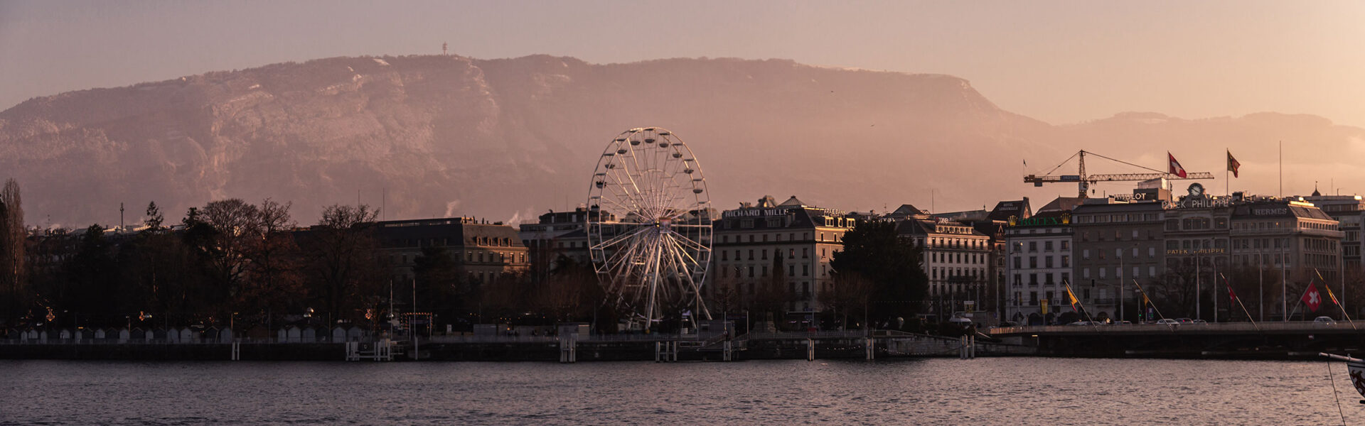 The shoreline in Geneva with a view of a ferris wheel, the city & mountains in the background.