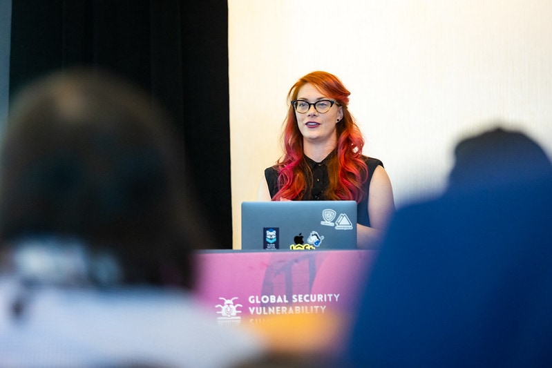 Woman with red hair and glasses addressing an audience at an event.