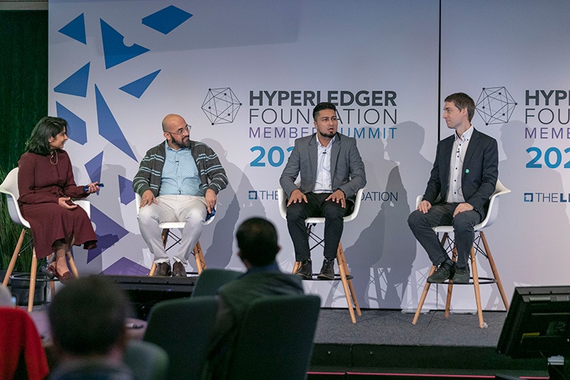 Four speakers sitting on chairs on stage with a sign behind them saying "Hyperledger Foundation Member Summit 2022; The Linux Foundation."