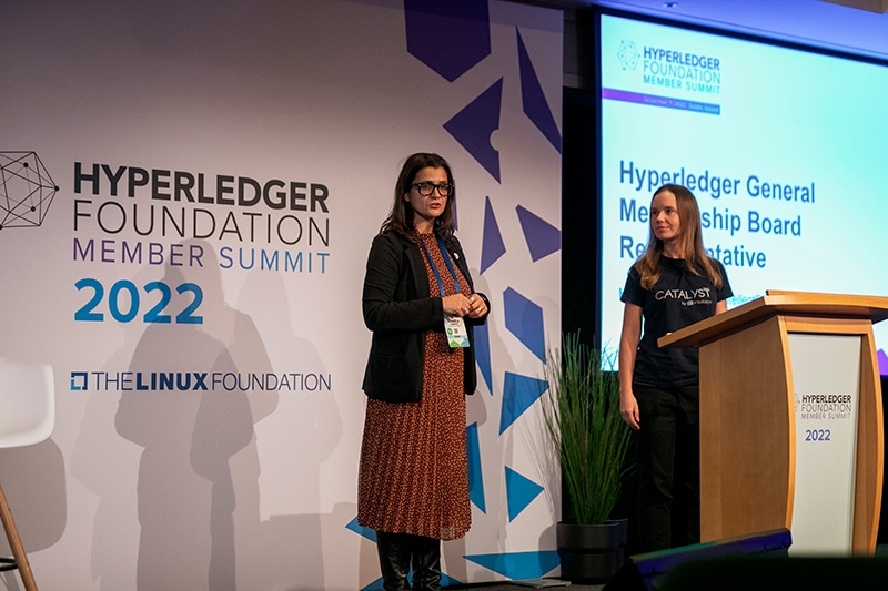 Two women speakers on stage addressing the crowd. There's a sign behind them that says "Hyperledger Foundation Member Summit 2022; The Linux Foundation."