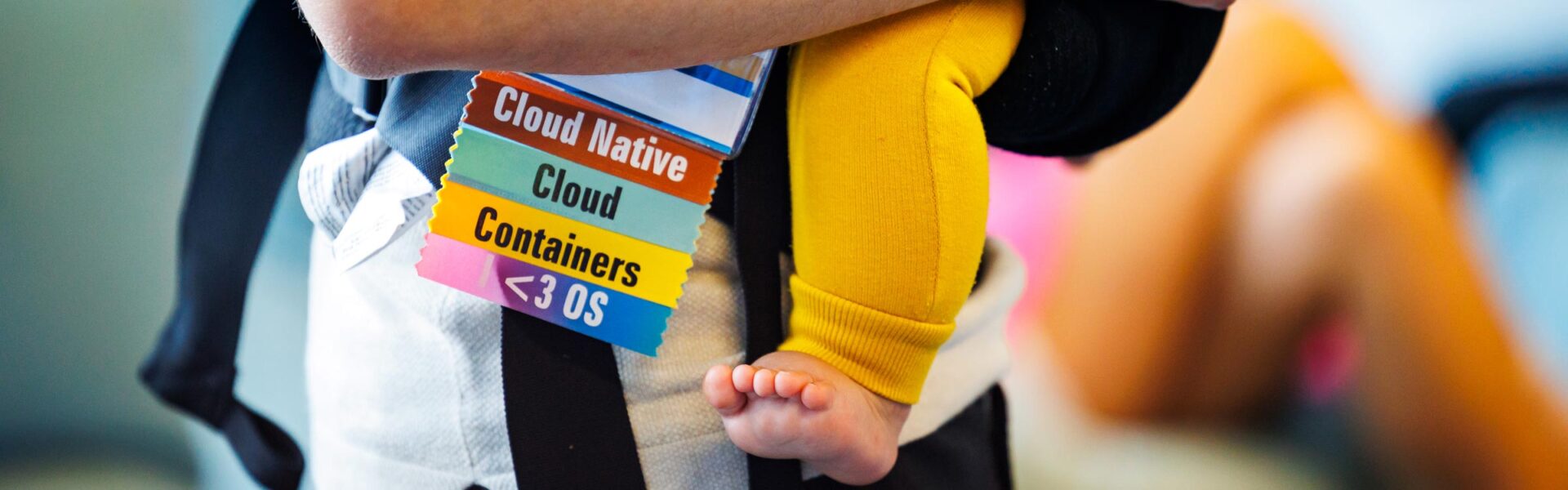 A parent holding a baby in a carrier. The adult has an event badge on with ribbons saying, "Cloud Native, Cloud, Containers, and I heart OS."