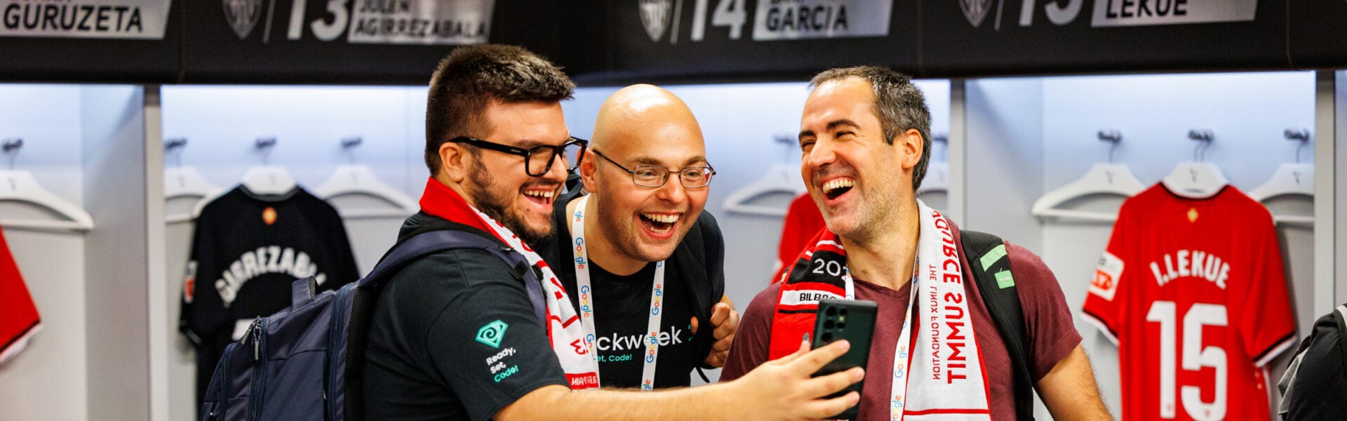 Three men laughing at something on a phone in a fútbol locker room, wearing Open Source Summit fútbol scarves.
