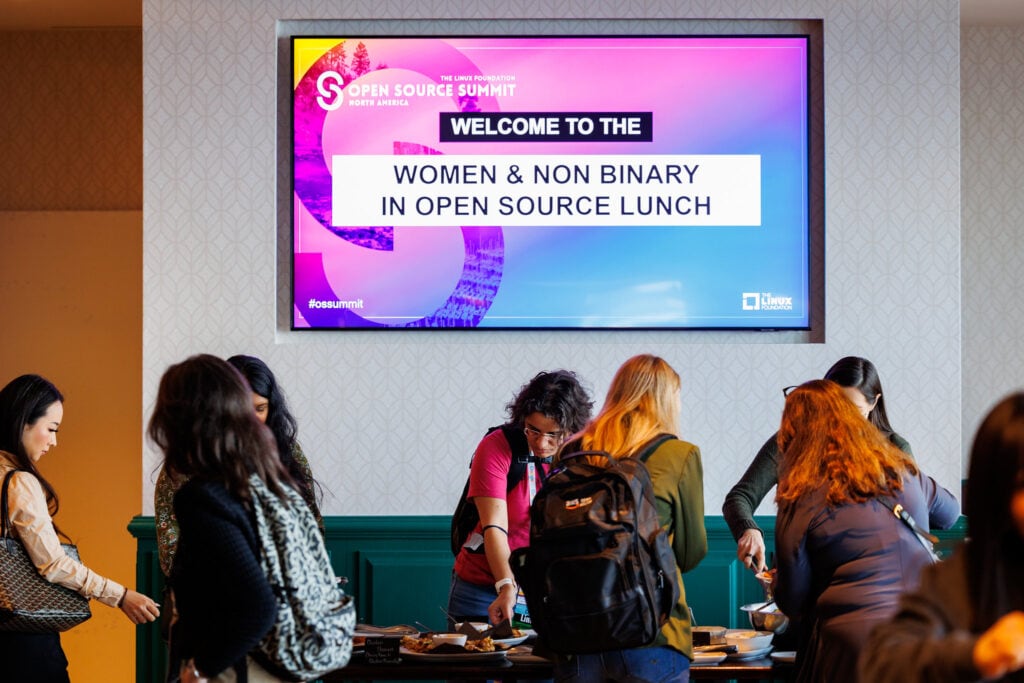 A sign that says "Welcome to the Women and Non Binary in Open Source Lunch" hang above a buffet lined with attendees.