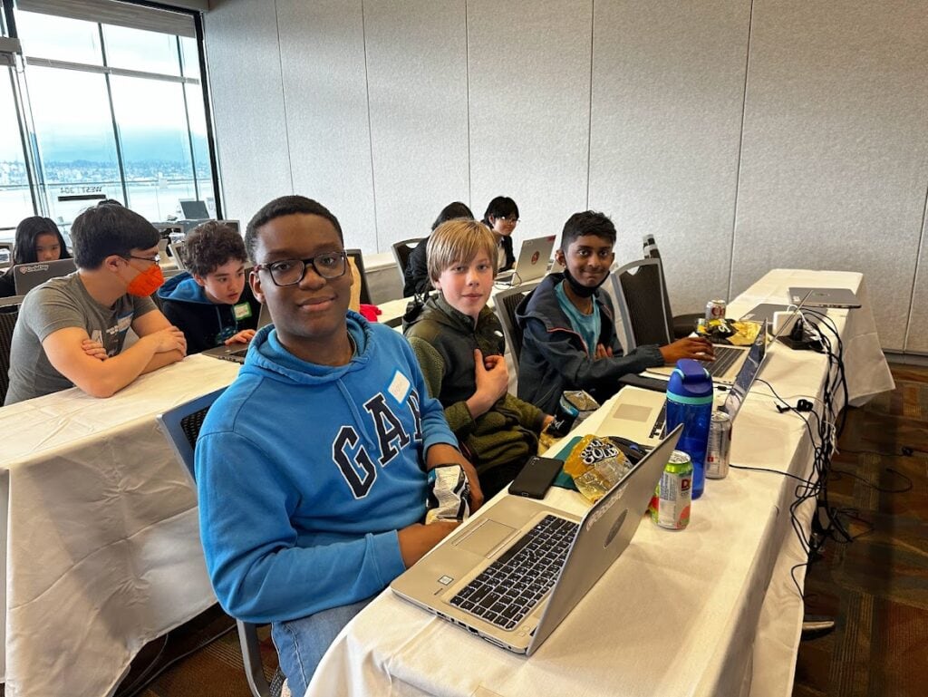 Three boys sit in front of their computers, smiling at the camera.