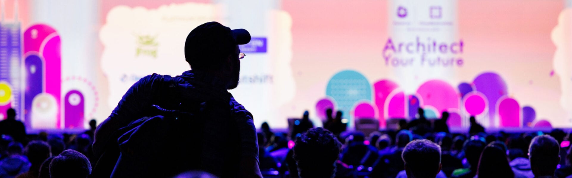 Silhouettes of the back of a seated crowd of attendees, with a man finding a seat for keynotes.