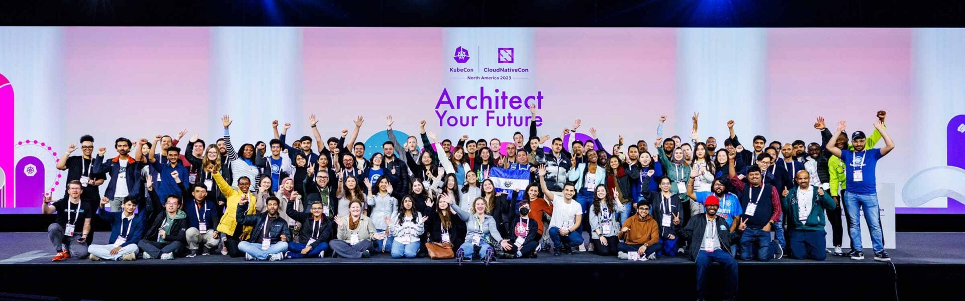 A large number of attendees, some with their arms raised, taking a group photo in front of a backdrop that says "Architect Your Future."