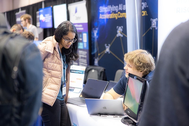 A woman looking at a laptop screen while talking to a man sitting behind a table.