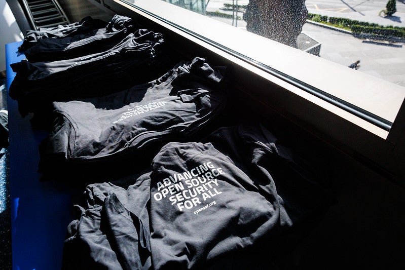 Stacks of black shirts that say "Advancing Open Source Security for All - openssf.org" underneath of a window.