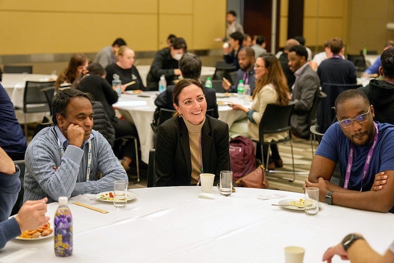 Attendees seated in a large room at round tables having snacks and talking.