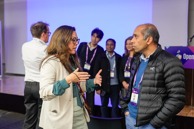 Two attendees talking to each other while a group poses for a photo behind them.