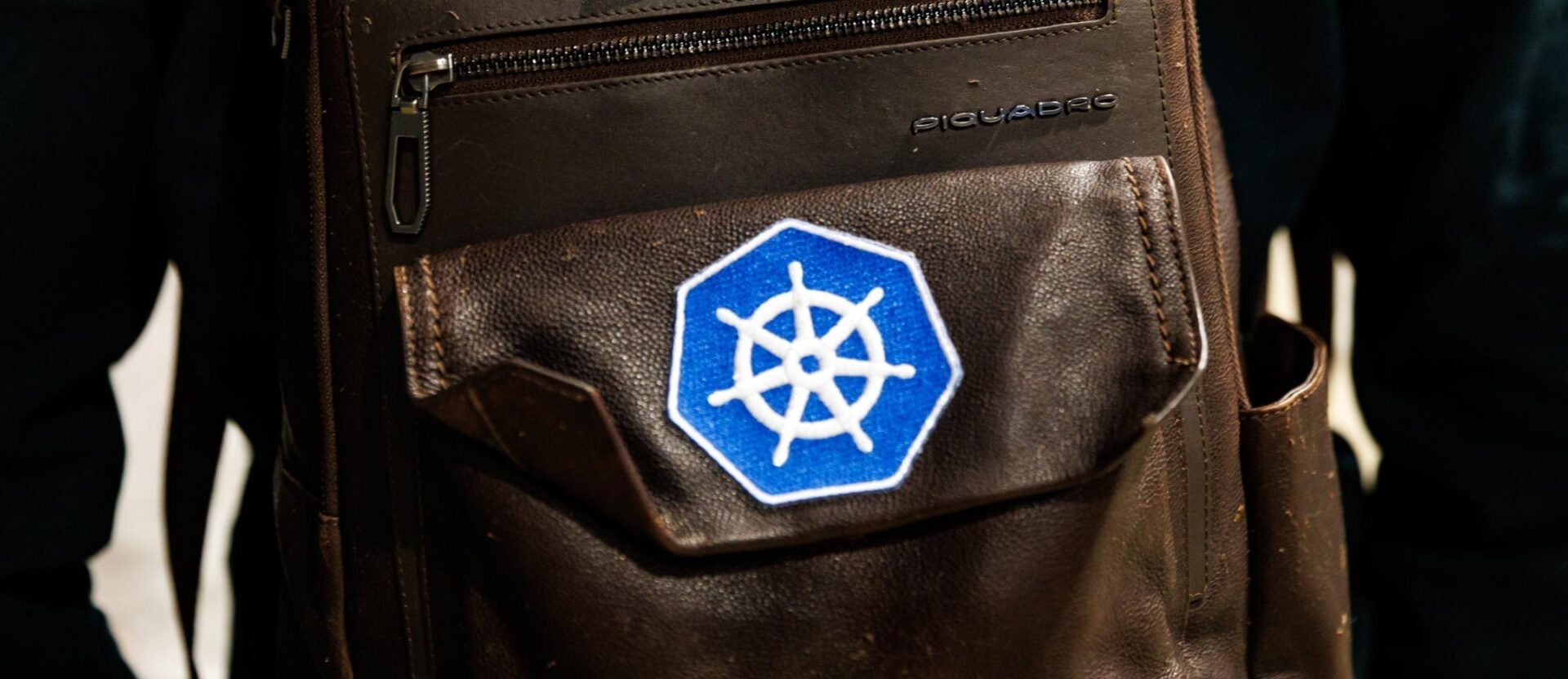 A brown backpack with a blue and white Kubernetes patch on it.