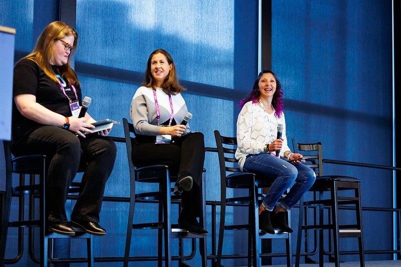 Three women smiling and laughing on a stage seated in tall chairs.