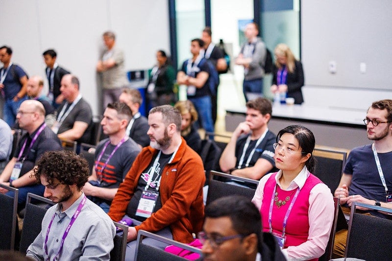 Attendees seated in rows and standing in the back of the room during a session.