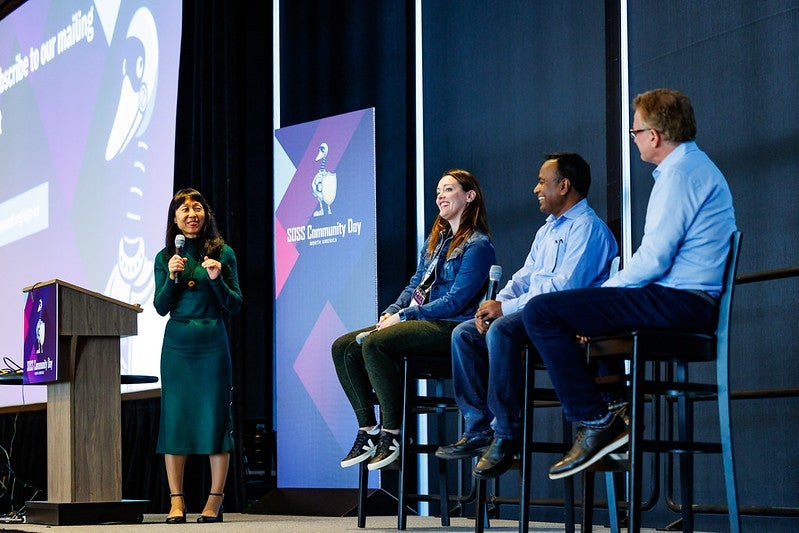 A woman standing near a podium with a microphone, talking with 3 panelists seated in tall chairs.