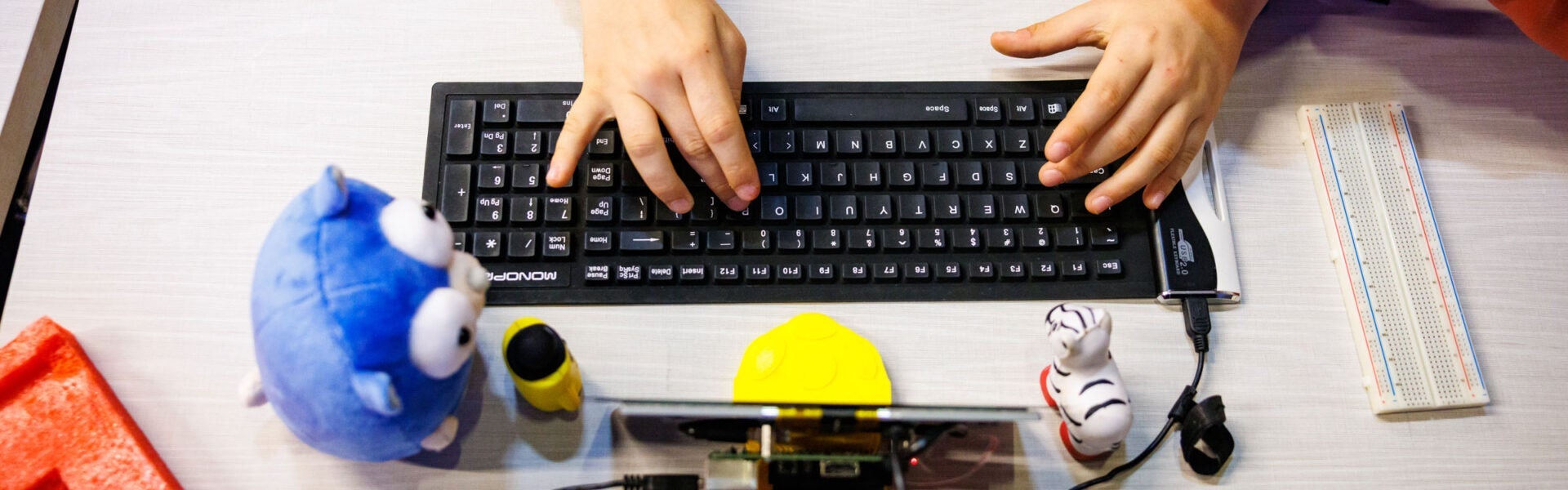 A childs hands on a computer keyboard with toys around it.