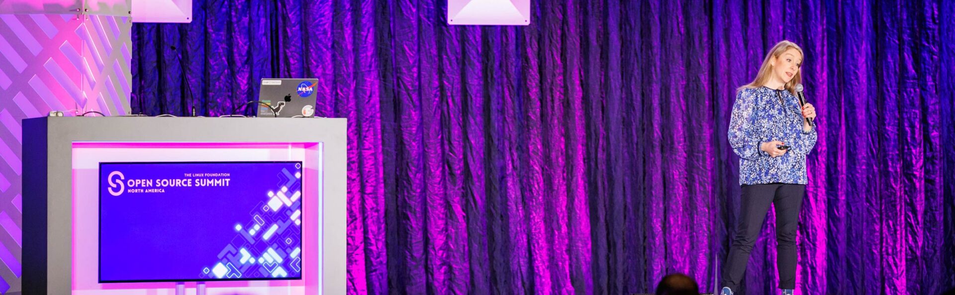 A woman on stage with a microphone addressing the crowd. There's a sign that says Open Source Summit North America on the podium.