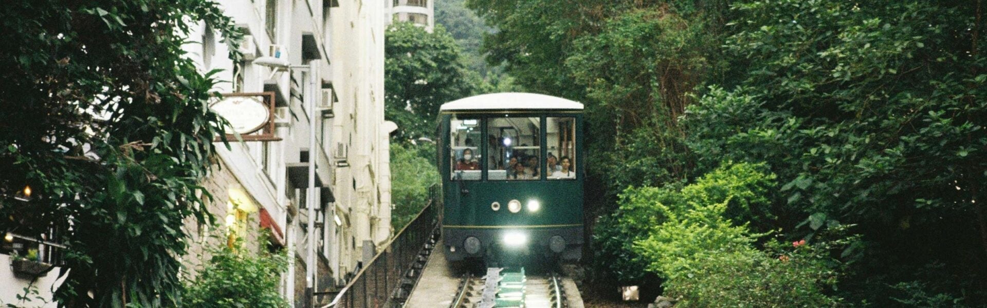 A green train on the tracks between buildings and a forest.