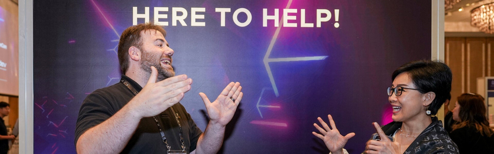 A sign that says "Here to Help!" with 2 attendees smiling at each other in front of it.