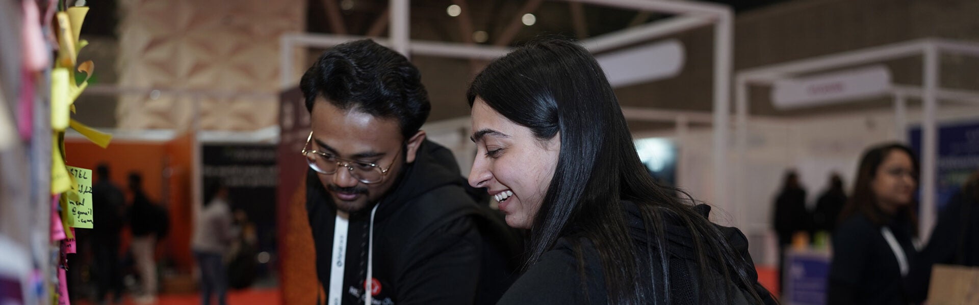 A man and a woman smiling and looking at the notes on a job board.