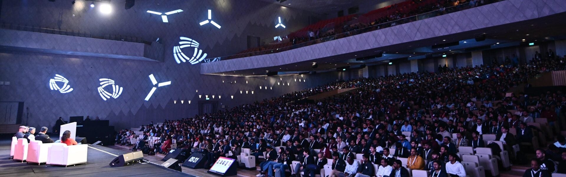 A wide angle view from the stage at an event showing the seated speakers & a full crowd of attendees.