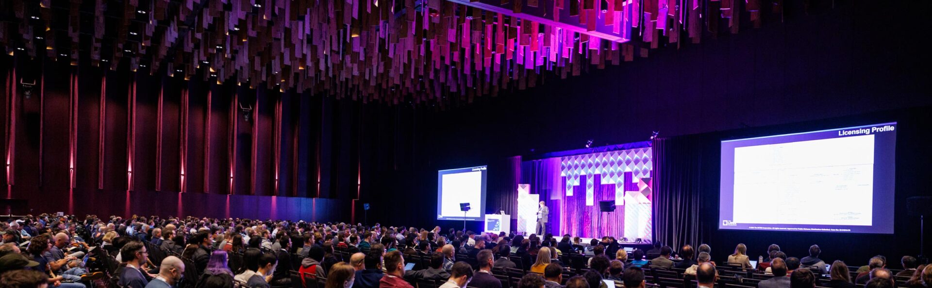 A view from the side of a keynote speaker room showing a large crowd & a speaker on stage.