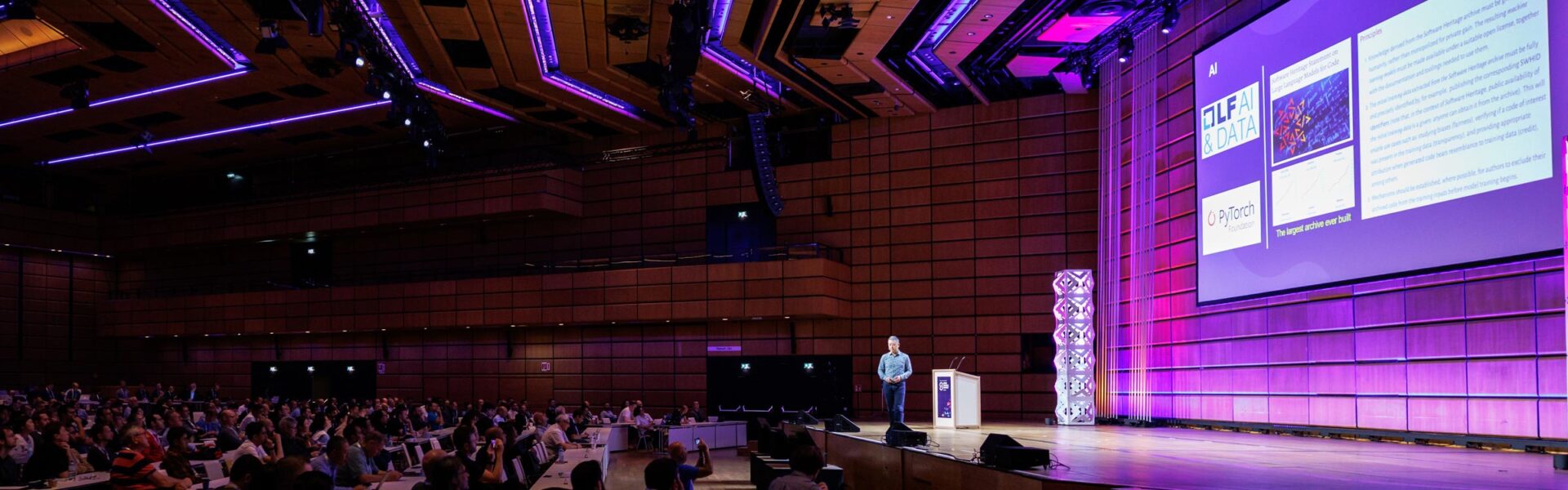 A view of a speaker on the keynote stage and a crowd with pink and purple lighting on the ceiling and stage.