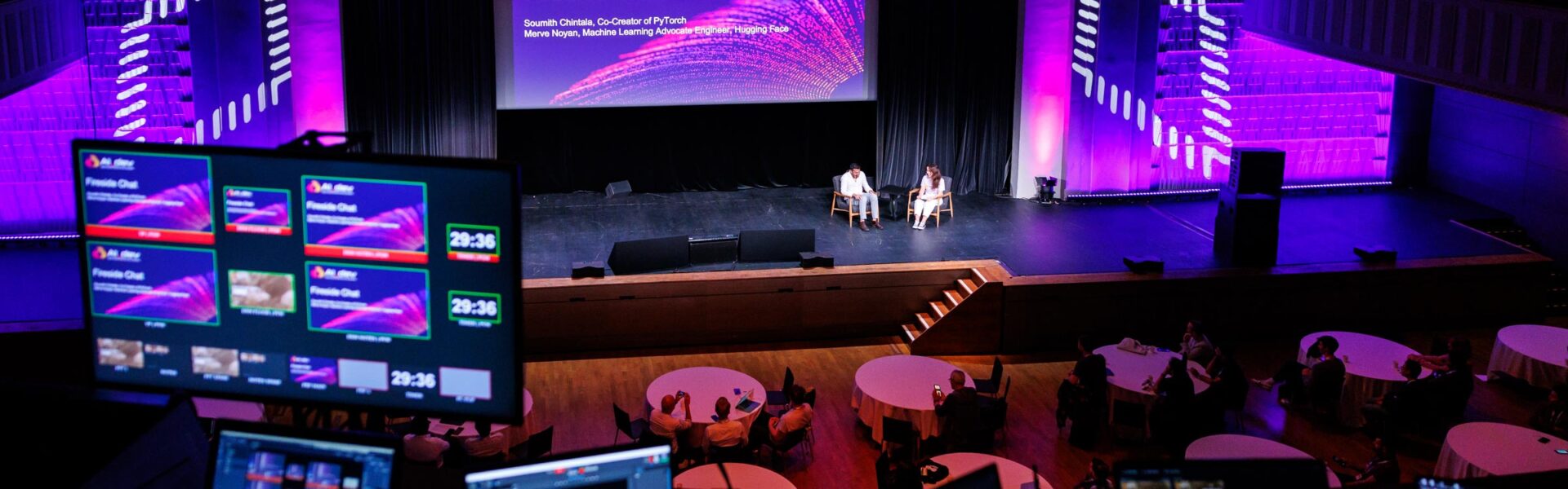 A high-angle view from the control area, showing two seated speakers on stage, a large screen, illuminated graphics, and close-up screens with slides and stage controls.