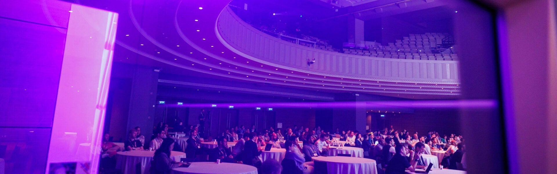 A large group of attendees seated at round tables in the keynote room, illuminated with pink and purple lighting.