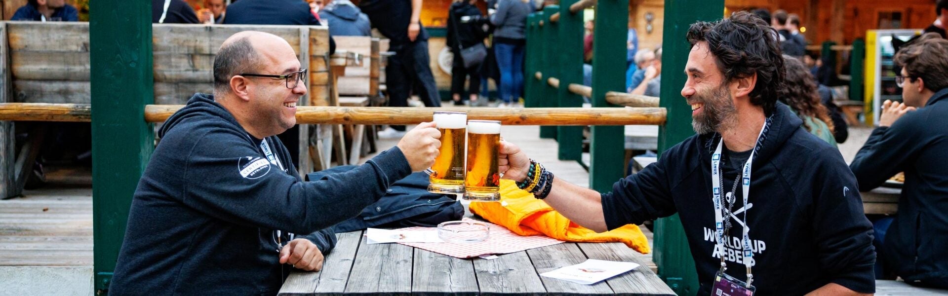 Two men seated outdoors at a wooden table clinking their beer glasses together in a toast.