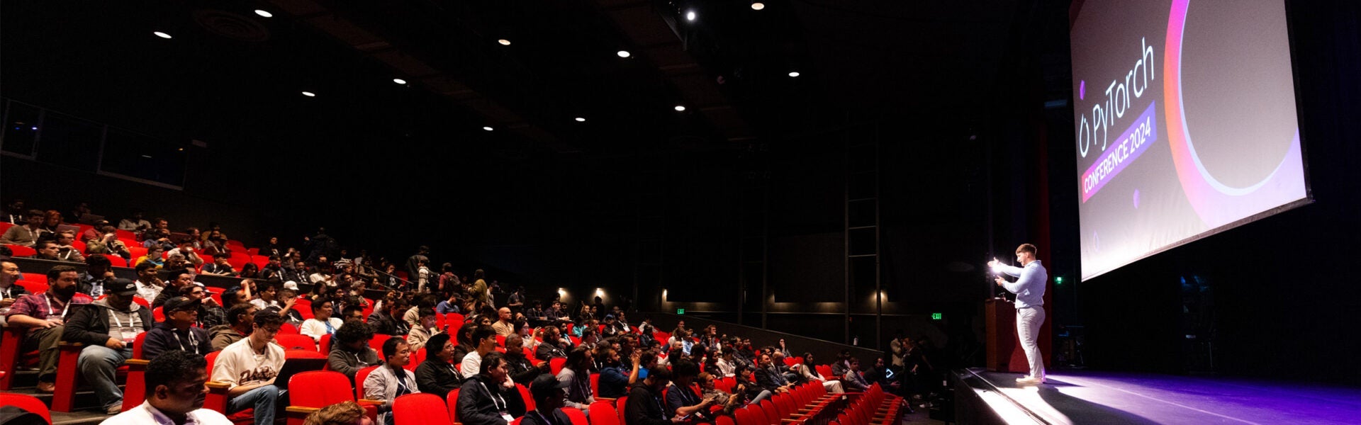 An auditorium filled with people listening to a speaker on stage.