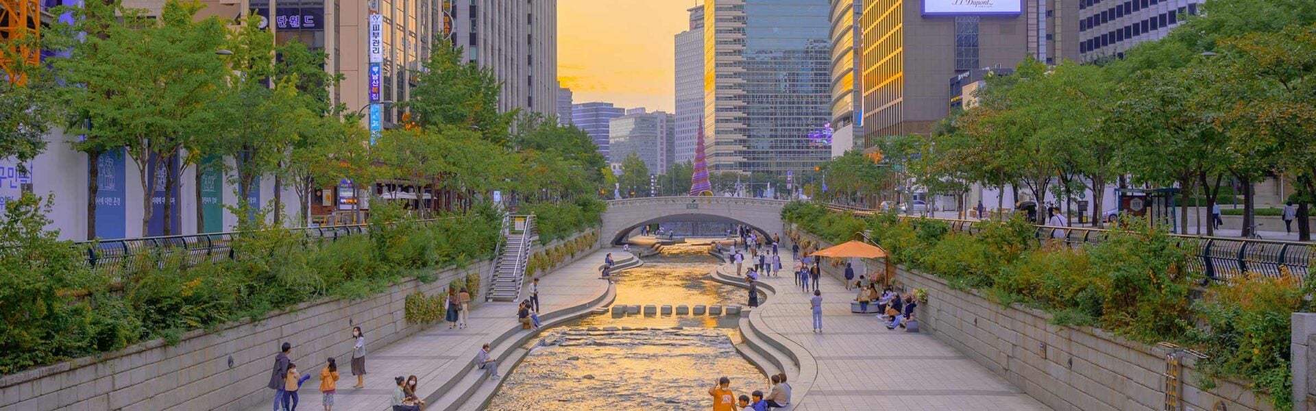 People walking and sitting on a sidewalk that lines a river in the city.