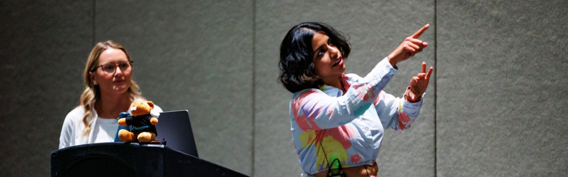 A woman in a colorful shirt speaking on stage and pointing up at a screen (that's out of view).