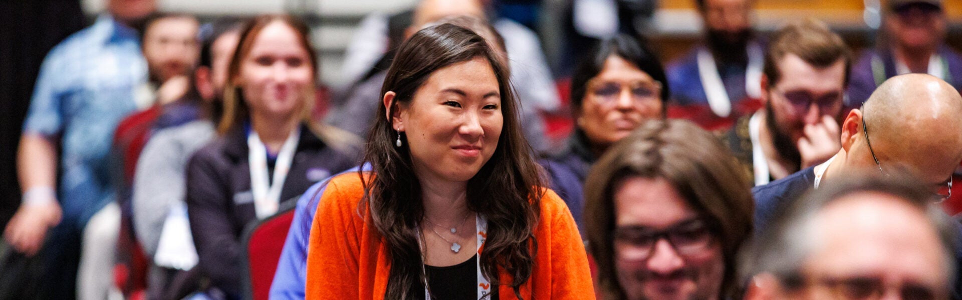 A woman in an orange sweater smiling with other audience members in the background.