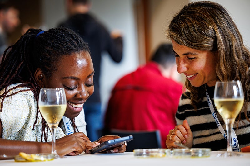 Two women seated at a table, smiling and looking at a phone with glasses of wine in front of them.
