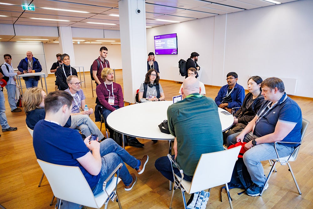 Event attendees seated & standing around a round white table having a discussion.