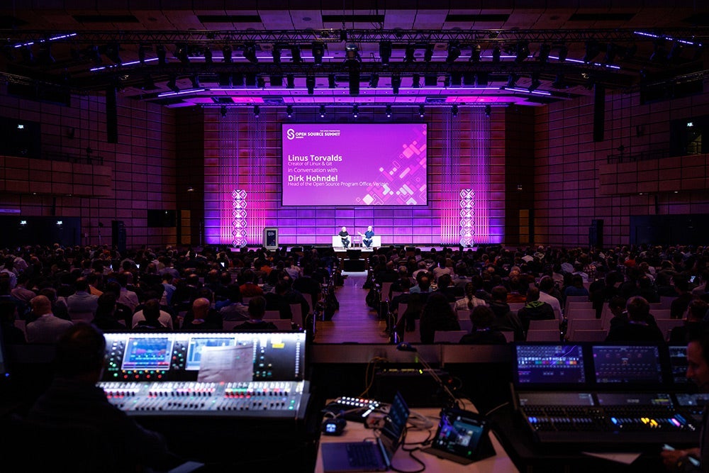 Two speakers seated on a brightly lit stage in a dimly lit keynote room.