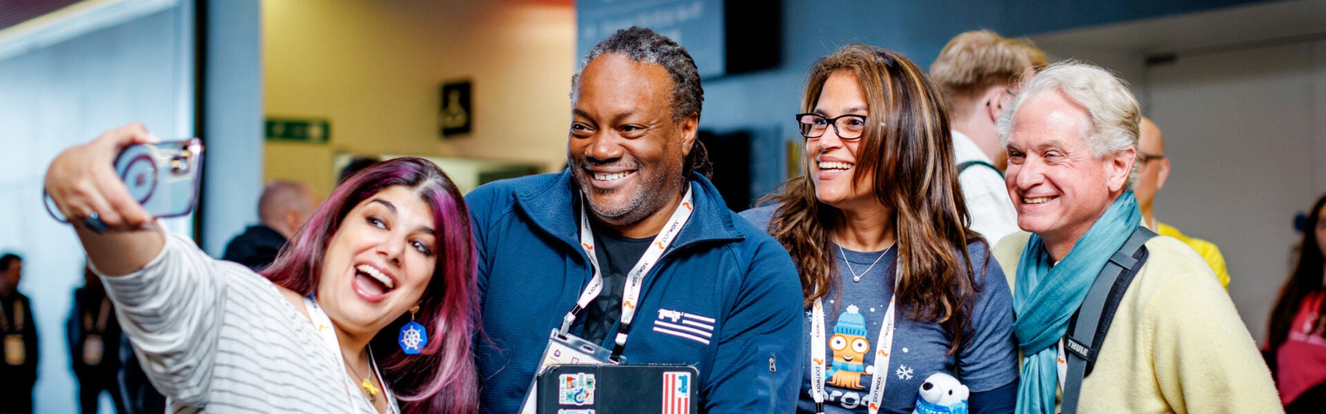Two women and two men smile for a selfie at an event. The woman taking the photo has purple hair and is wearing Kubernetes earrings.