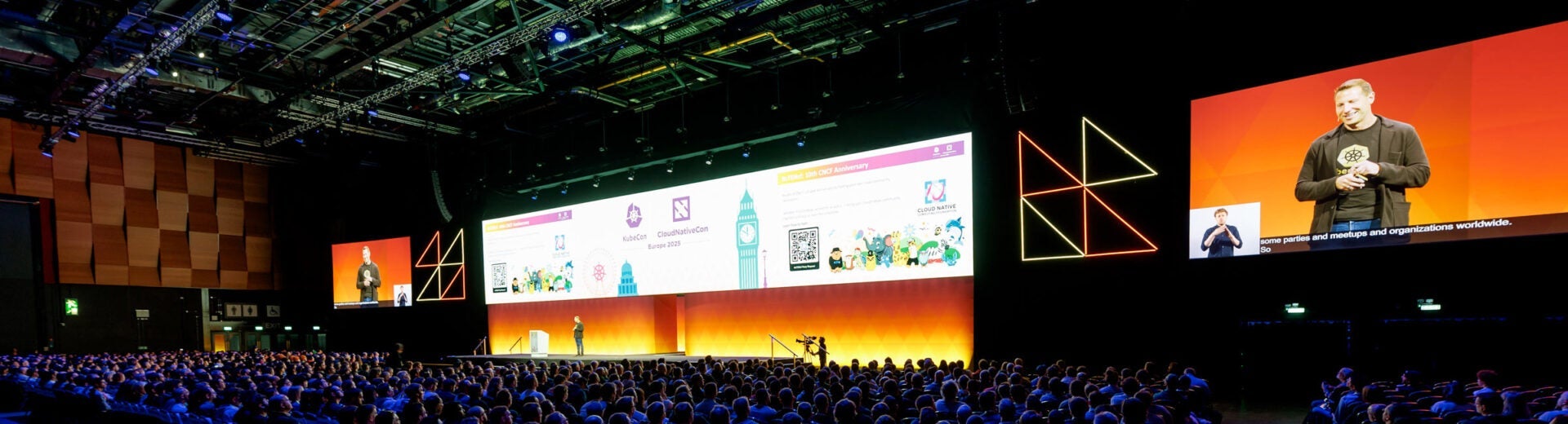 A keynote speaker presents on stage in a large, dimly lit auditorium. The presentation screens display the speaker, live captioning, and a sign language interpreter.