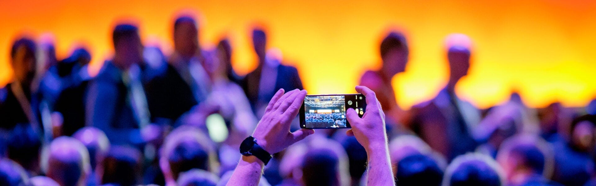 A person in the audience holds up a phone to take a photo in a large event space, where attendees are seated in rows and others are standing and finding their seats.