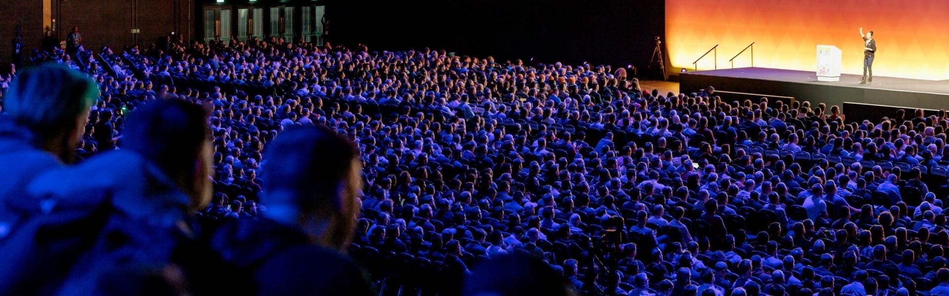 A keynote speaker presents on stage to a large audience in a dimly lit auditorium with blue and orange lighting.