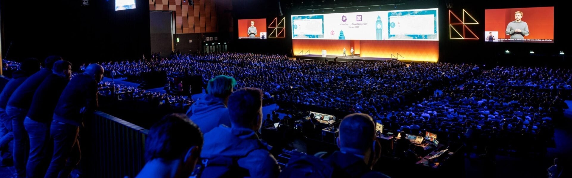 Large crowd seated in a dark auditorium during the KubeCon + CloudNativeCon Europe 2025 keynote. Two speakers are on stage in front of a wide, brightly lit screen with event branding. A live video feed of one speaker is displayed on a large screen to the right, alongside colorful geometric lighting.