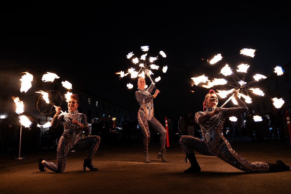 Three fire dancers performing outdoors on a concrete surface at night, each striking a pose while twirling flaming props.