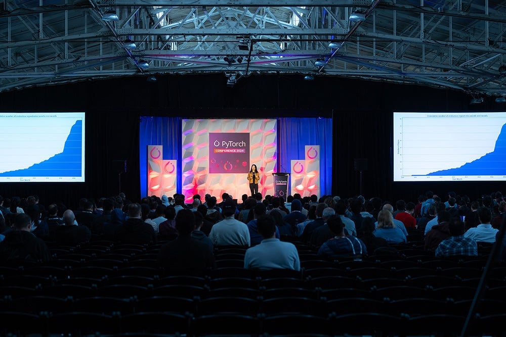 Speaker onstage at PyTorch Conference 2024, with a colorful display behind them, viewed from the back of a large conference room filled with attendees.