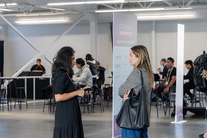 Two women talking in a room filled with people.