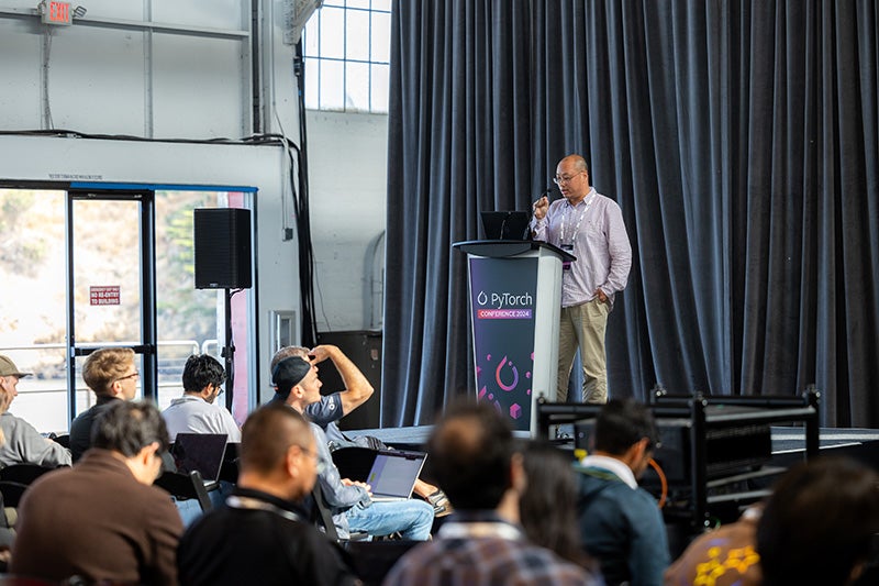 A speaker at a podium on stage addressing a room of people.